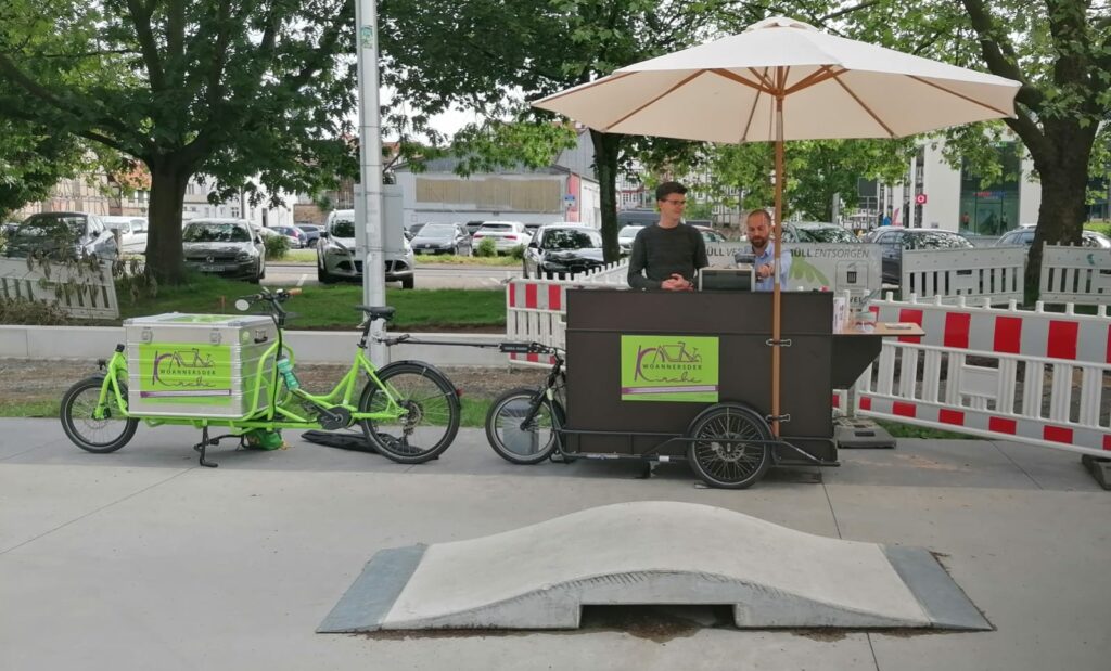 Ein grünes Lastenrad, vorne eine Metallkiste, hinten ein Anhänger, der zu einer mobilen Kaffeebar umgebaut werden kann. Im Vordergrund eine kleine Skaterampe.