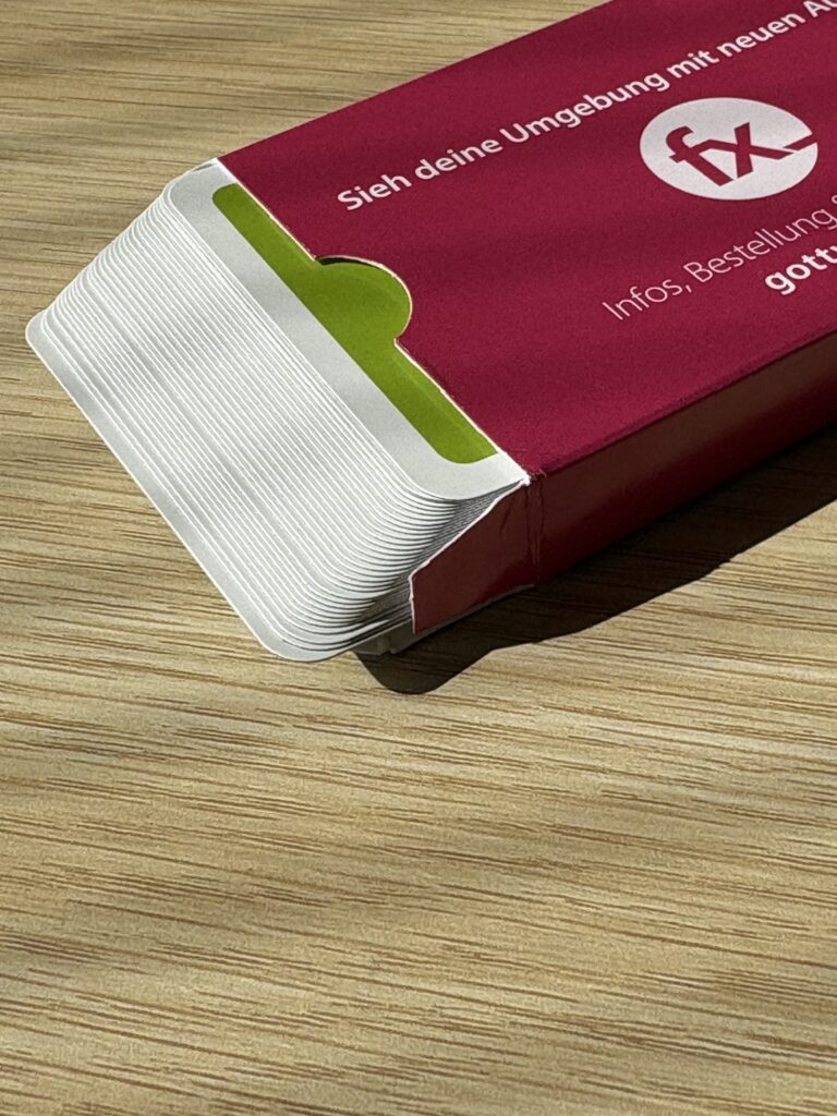 Eine teilweise geöffnete rot-weiße Pappschachtel liegt auf einer hölzernen Unterlage und gibt den Blick auf weiße Karten im Inneren frei. Das Sonnenlicht wirft Schatten auf die Schachtel und den Tisch.