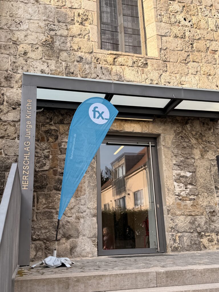 Eine blaue Fahne steht vor einem steinernen Gebäude mit einer Glastür. Auf dem Schild steht HERZSCHLAG Junge Kirche. Ein Fenster spiegelt nahe gelegene Gebäude.