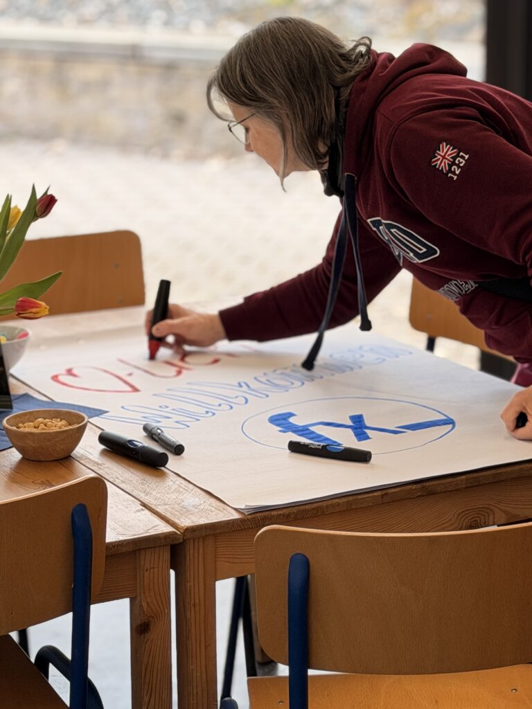 Eine Person in einem kastanienbraunen Kapuzenpulli schreibt mit Stiften auf einem großen Blatt Papier an einem Tisch mit Stühlen, Tulpen und einer Schale mit Snacks in der Nähe.