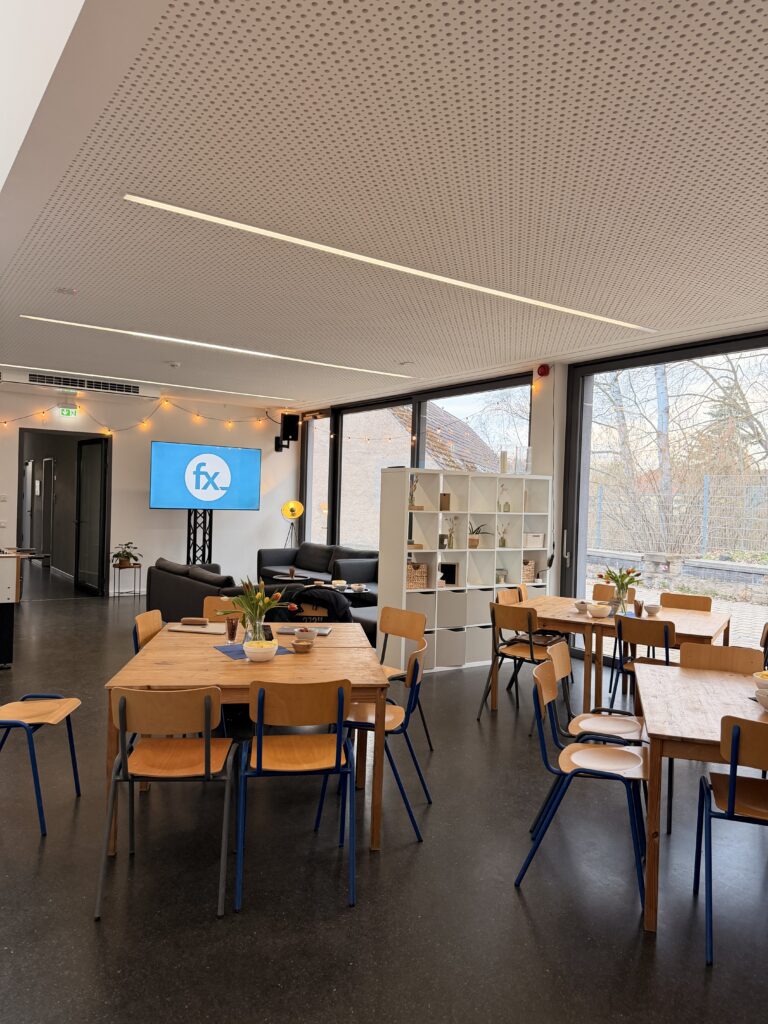 Ein moderner Lounge-Bereich mit Holztischen und -stühlen, Topfpflanzen, Sofas, einem Fernseher mit Filmen, großen Fenstern und Regalen mit Dekorationsartikeln.