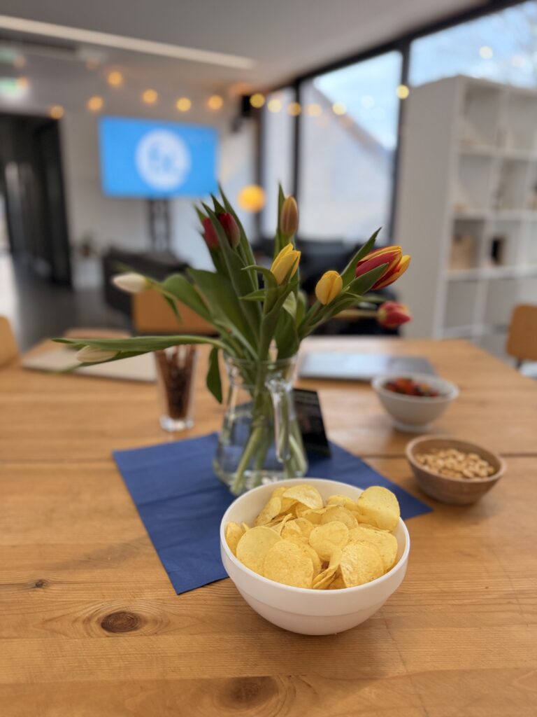 Eine Schale mit Kartoffelchips auf einem Holztisch mit einer Vase mit Tulpen, Snacks und einem unscharfen Bildschirm im Hintergrund.