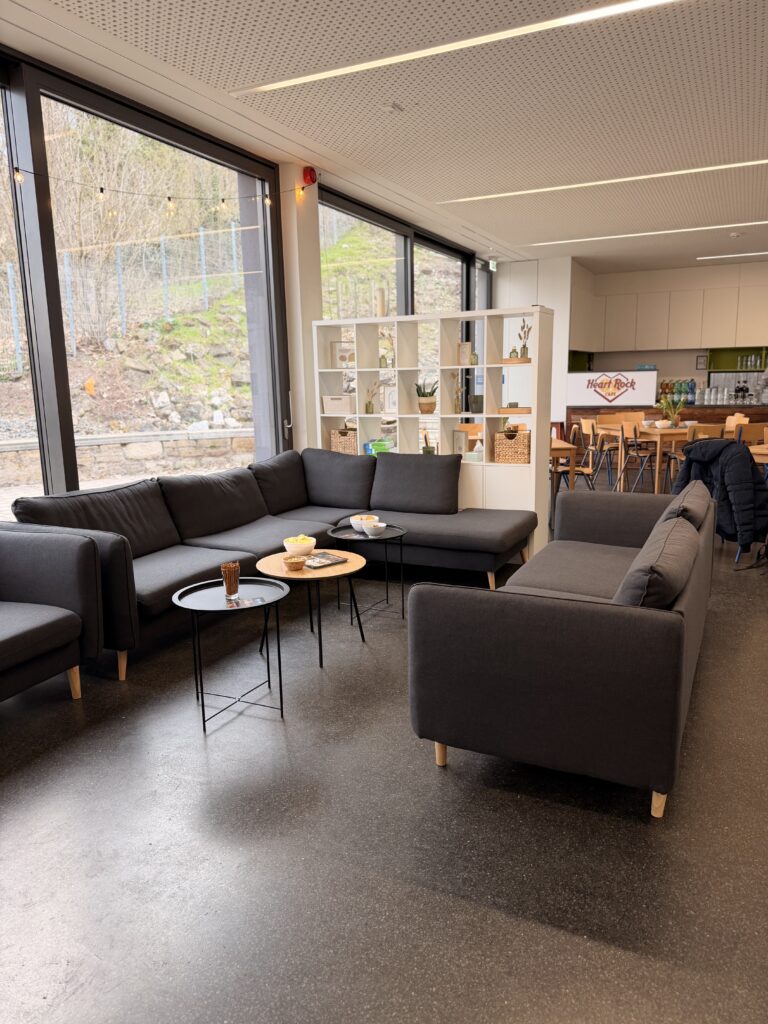 Moderner Lounge-Bereich mit schwarzen Sofas, kleinen runden Tischen mit Snacks und Getränken, großen Fenstern, einem weißen Bücherregal und einem Küchenbereich im Hintergrund.