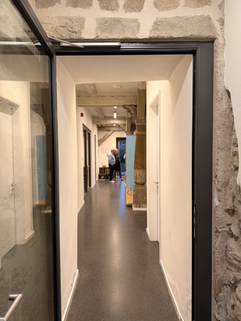 Ein Flur mit steinernen und weißen Wänden, einer offenen Glastür und einer Person, die in der Ferne in der Nähe von Kisten geht. Der Boden ist dunkel und glänzend.
