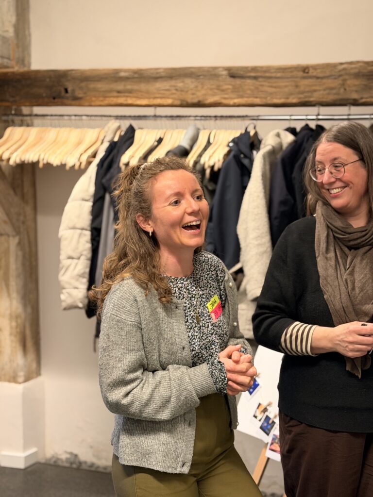 Zwei Frauen lächeln und unterhalten sich, während sie in einem Haus stehen. Hinter ihnen hängen Mäntel auf einem Gestell und an der Wand ist ein Holzbalken zu sehen.