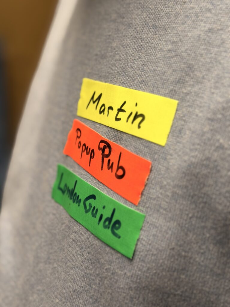 Drei farbige Etiketten auf einem grauen Stoff lauten: Martin (gelb), Popup Pub (orange) und London Guide (grün), jeweils handgeschrieben mit schwarzem Marker.