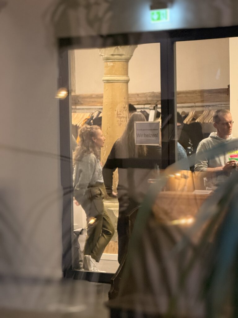 Drei Personen sind durch eine Glastür in einem warm beleuchteten Raum zu sehen, mit Holzdekor und aufgehängter Kleidung im Hintergrund.