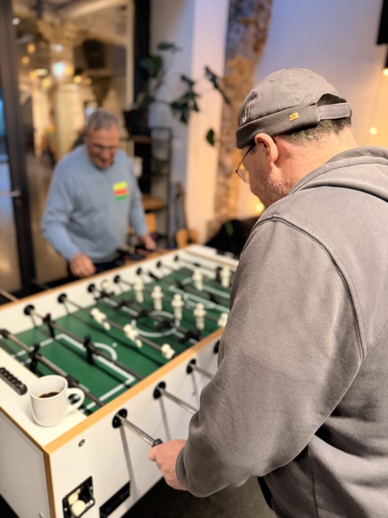 Zwei Personen spielen in einem Haus Tischfußball. Einer trägt im Vordergrund einen grauen Kapuzenpulli, während der andere am Tisch steht. Ein Kaffeebecher steht auf dem Tisch.