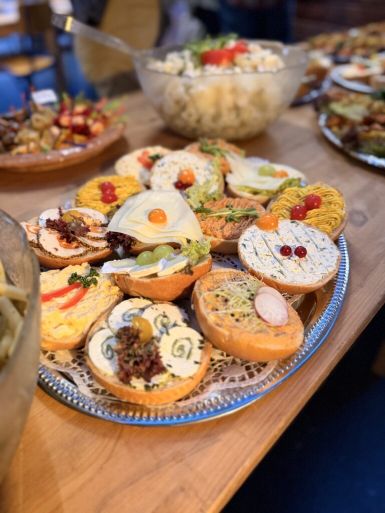Eine Platte mit verschiedenen belegten Brötchen, umgeben von anderen Speisen auf einem Holztisch an einem Buffet.