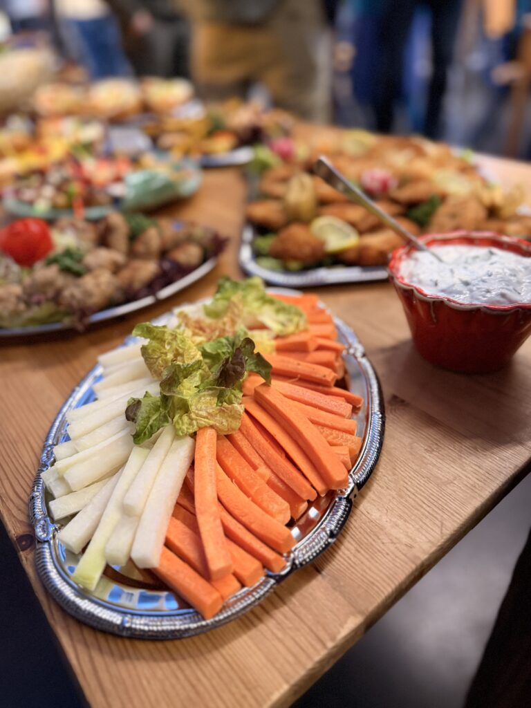 Ein Teller mit Karotten- und Jicamascheiben mit Salatgarnitur steht auf einem Holztisch neben einer Schüssel mit Dip und Tabletts mit verschiedenen Vorspeisen im Hintergrund.