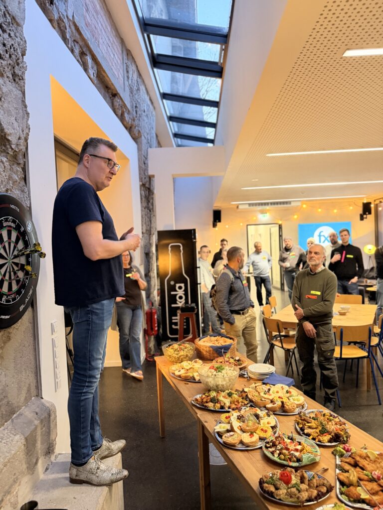 Ein Mann steht auf einer Stufe und wendet sich an eine Gruppe in einem Innenraum, während im Vordergrund ein Tisch mit verschiedenen Speisen steht und im Hintergrund Menschen zuhören.