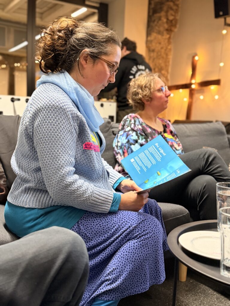 Zwei Frauen sitzen auf einer Couch in einem Innenraum; eine hält ein buntes Heft in der Hand und liest darin, während die andere nach vorne schaut. Gläser und ein Tablett stehen auf einem Tisch im Vordergrund.