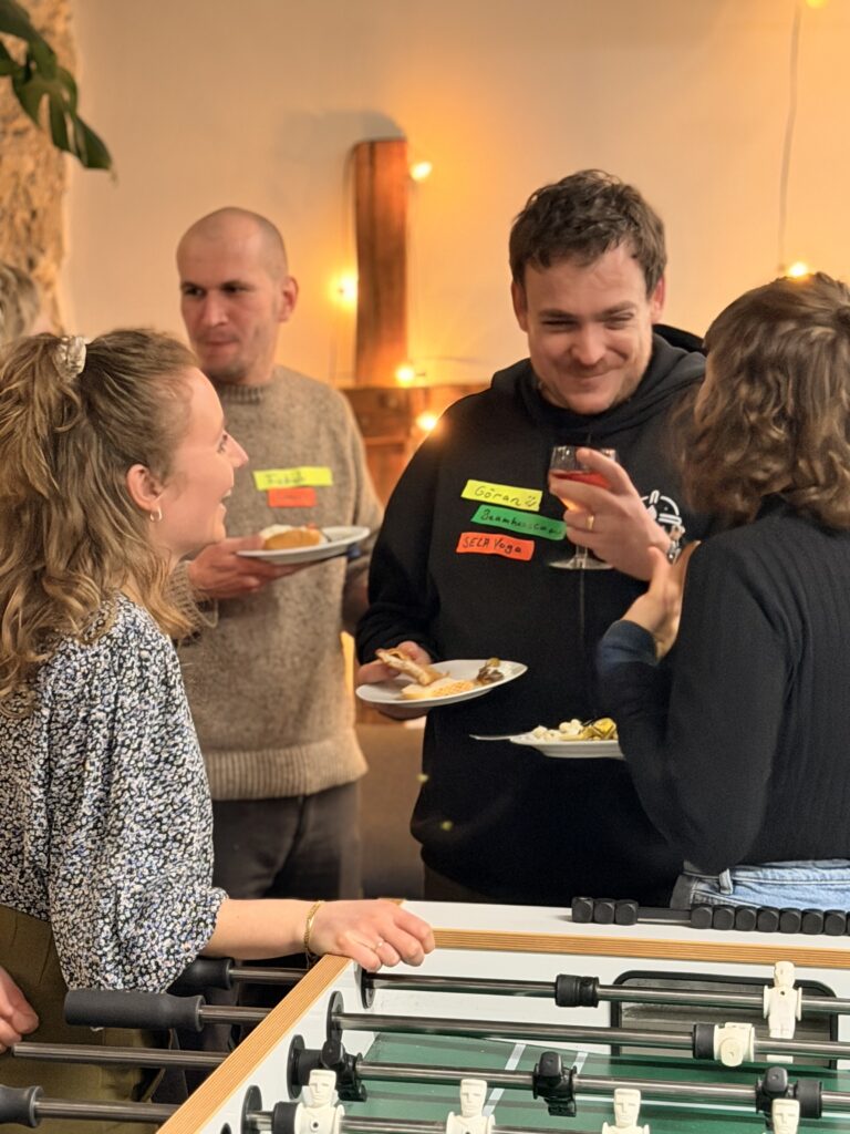 Vier Personen stehen bei einem zwanglosen Treffen um einen Kickertisch herum, halten Teller mit Essen und Getränken in der Hand, plaudern und lächeln mit Namensschildern an der Kleidung.