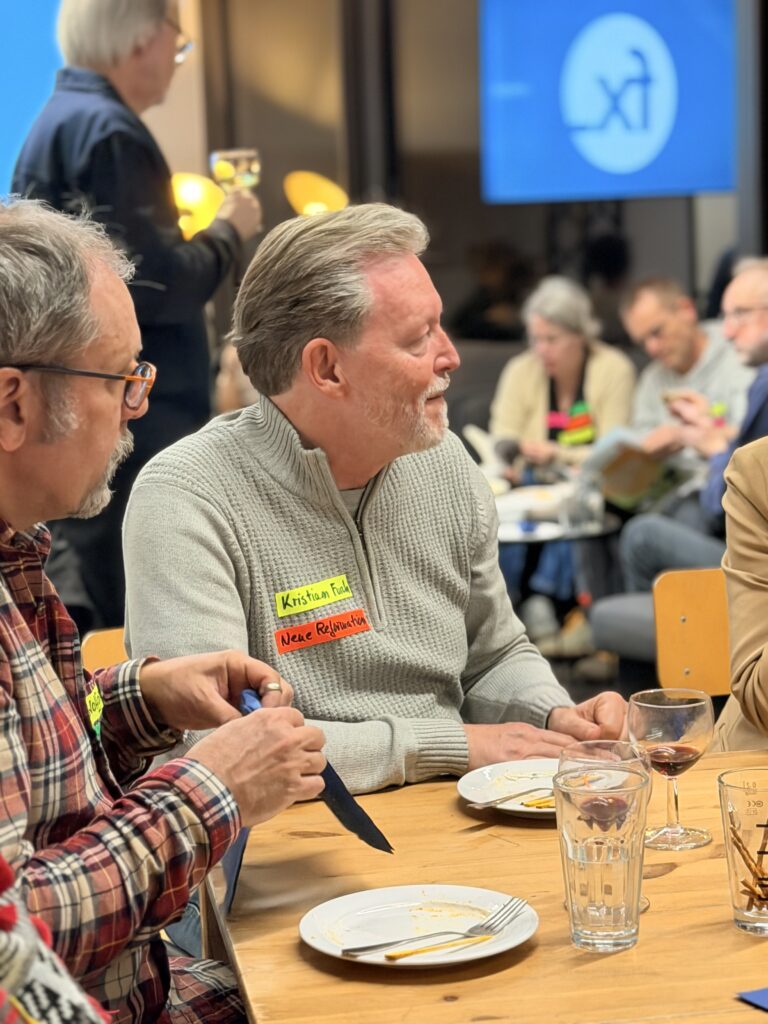 Zwei Männer sitzen an einem Tisch mit leeren Tellern und Getränken, mit Namensschildern, bei einem geselligen Beisammensein mit Menschen im Hintergrund.