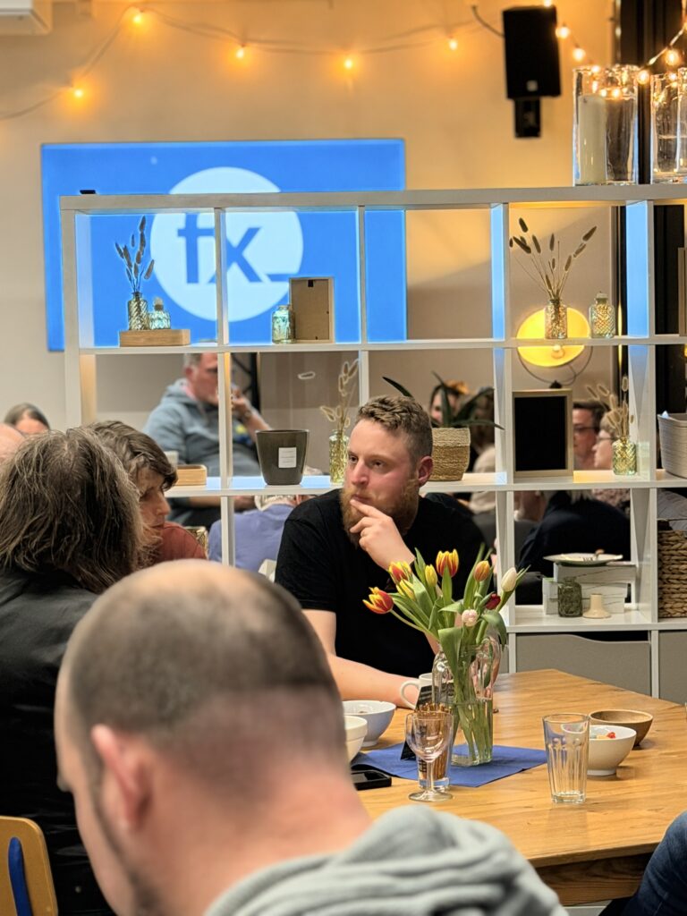 Menschen sitzen an Tischen in einem gemütlichen Café mit Lichterketten und Regalen, unterhalten sich und essen; ein Mann mit Bart steht im Mittelpunkt neben einer Vase mit Tulpen.