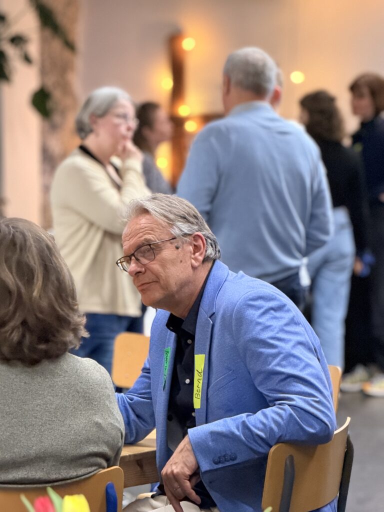 Ein Mann in einem blauen Blazer mit einem Namensschild sitzt an einem Tisch und spricht mit einer Frau; im unscharfen Hintergrund stehen mehrere Personen und unterhalten sich.