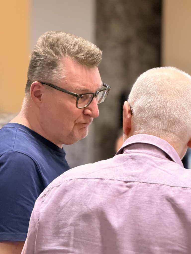 Zwei Männer unterhalten sich in einem Haus. Der eine Mann blickt in die Kamera, trägt eine Brille und ein blaues Hemd, der andere hat kurzes weißes Haar und trägt ein hellviolettes Hemd.