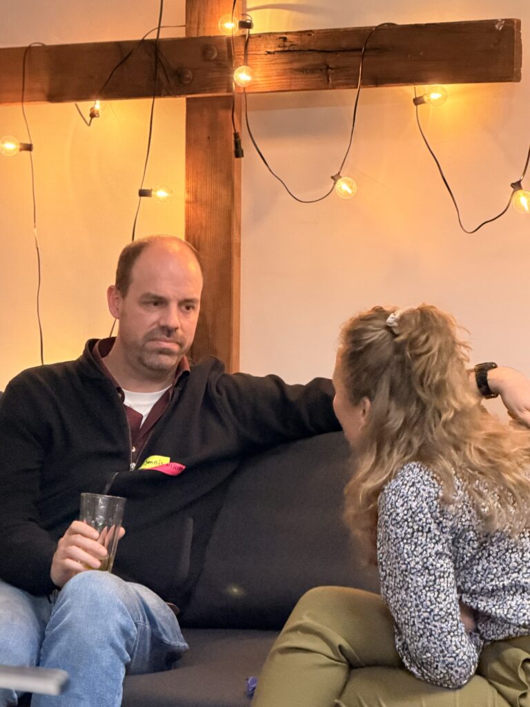 Ein Mann mit einem Glas in der Hand sitzt mit ernster Miene auf einer Couch und spricht mit einer Frau mit langen Haaren vor einem Holzkreuz mit Lichterketten.