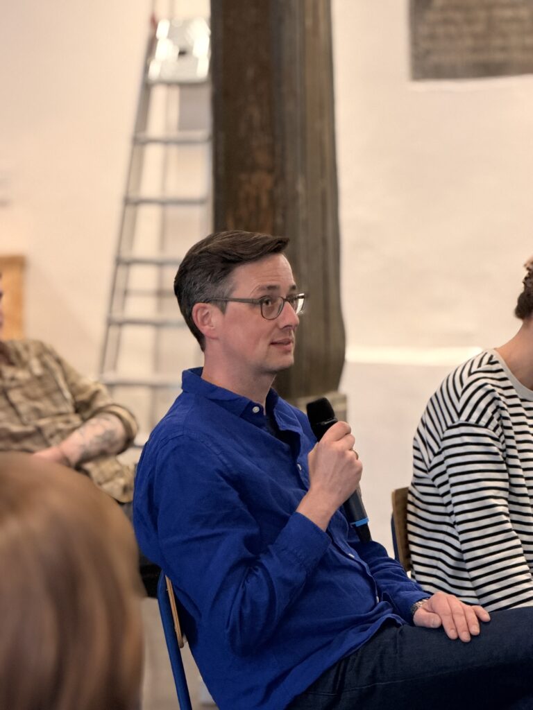 Ein Mann in einem blauen Hemd sitzt und spricht während einer Veranstaltung in einer Halle in ein Mikrofon, während andere Personen in der Nähe sitzen und eine Leiter im Hintergrund zu sehen ist.