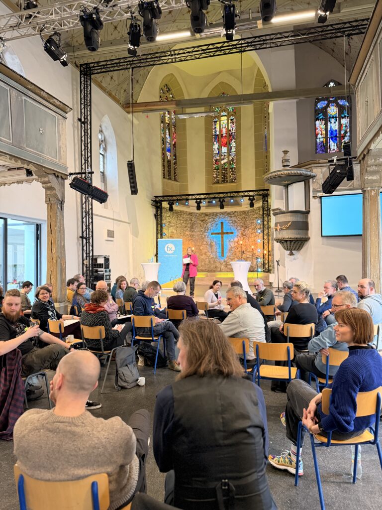 Eine Gruppe von Menschen sitzt in einem Kirchensaal mit Buntglasfenstern und hört einem Redner auf der Bühne neben einem blauen Kreuz und Präsentationsbildschirmen zu.