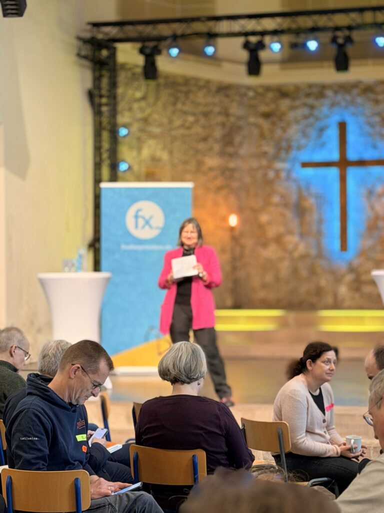 Eine Frau steht auf der Bühne und hält Papiere vor einem Hintergrund mit einem blauen Kreuz in der Hand, während im Vordergrund sitzende Personen zuhören und sich Notizen machen, während eine Veranstaltung in einer Halle stattfindet.