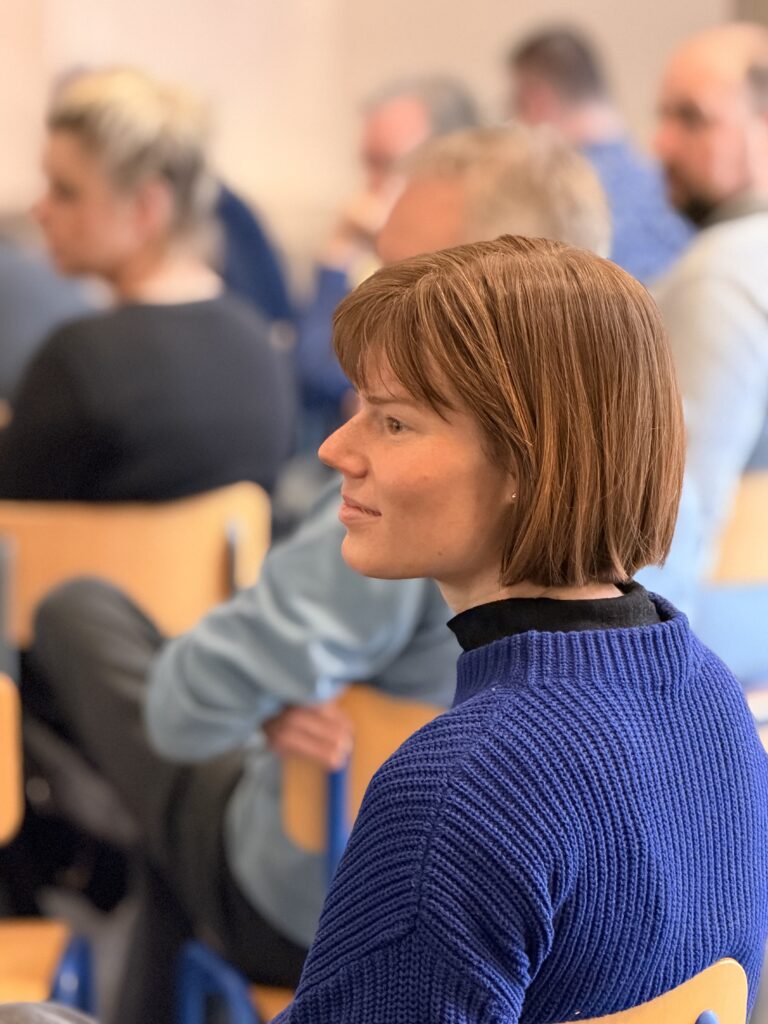 Eine Frau in einem blauen Pullover sitzt während einer Gruppensitzung oder eines Seminars und hört aufmerksam zu, während andere Personen im Hintergrund sitzen.