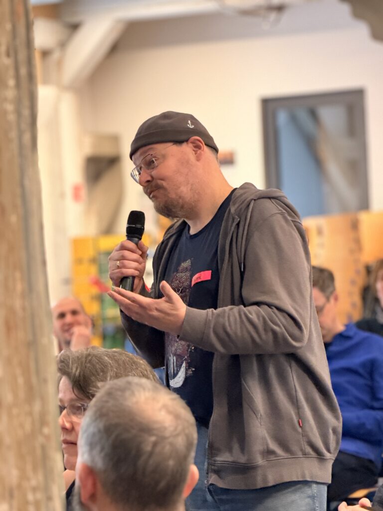 Ein Mann mit grauem Kapuzenpulli und Mütze spricht in ein Mikrofon, während er inmitten eines sitzenden Publikums in einem hell erleuchteten Innenraum steht.