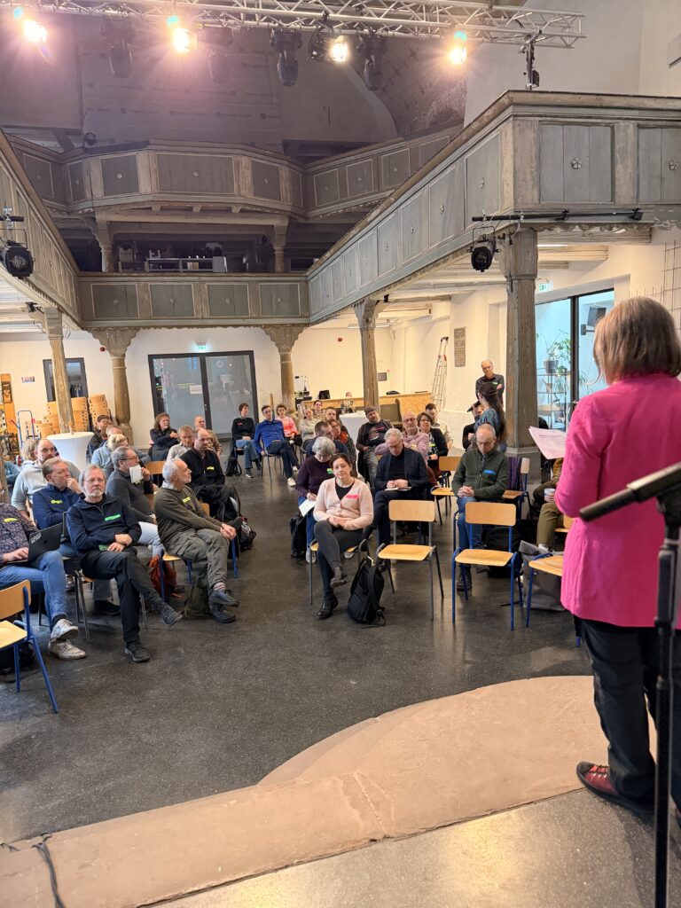 Eine Frau in einer rosafarbenen Jacke spricht zu einem Publikum, das auf Stühlen in einem geräumigen, rustikalen Saal mit Holzbalkonen und hohen Decken sitzt.