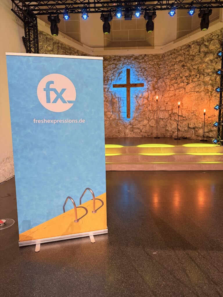 Ein freshexpressions.de-Banner steht in einer Kirche mit einem beleuchteten Kreuz an einer Steinwand und leuchtenden gelben Lichtern auf den Stufen dahinter.