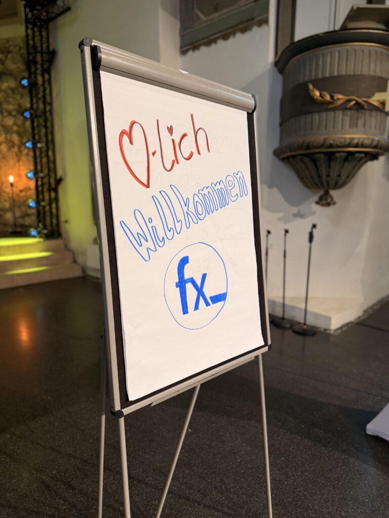 Auf einem Flipchart steht "Herzlich Willkommen" mit einem Herz und einer Grafik. Die Szene befindet sich in einem Innenraum mit Bühnenbeleuchtung und einer großen Wandleuchte im Hintergrund.
