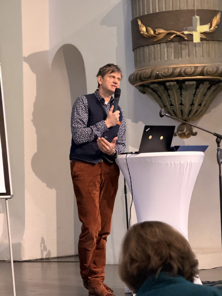 Ein Mann in einem gemusterten Hemd und einer braunen Hose spricht in ein Mikrofon, während er neben einem Laptop auf einem hohen runden Tisch bei einer Indoor-Veranstaltung steht.