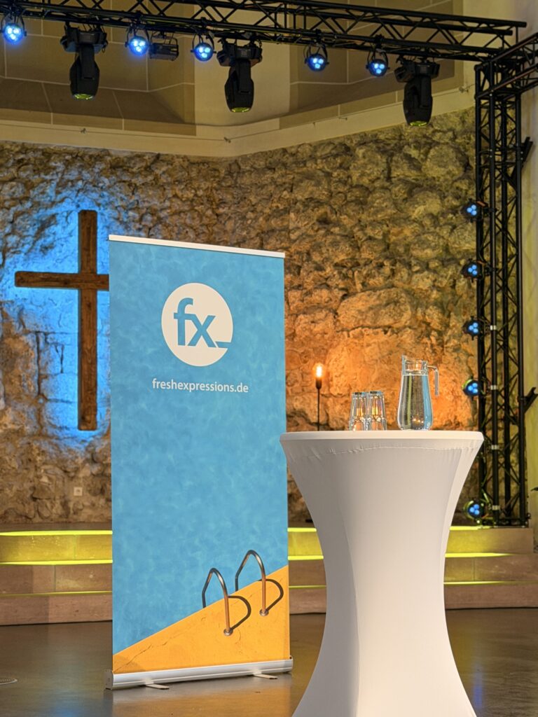 In einer Kirche mit Steinmauern, einem Kreuz und blauer und orangefarbener Beleuchtung steht ein runder Tisch mit Wasserkrügen neben einem FX-Banner.