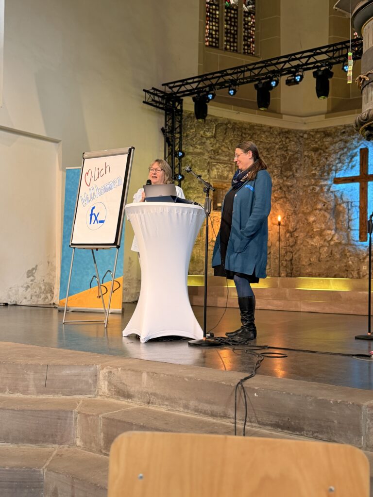 Zwei Frauen stehen an einem Podium in einer Kirche und sprechen. Auf einem Flipchart neben ihnen steht Herzlich Willkommen fx. Die Bühne ist beleuchtet und hat ein Kreuz im Hintergrund.