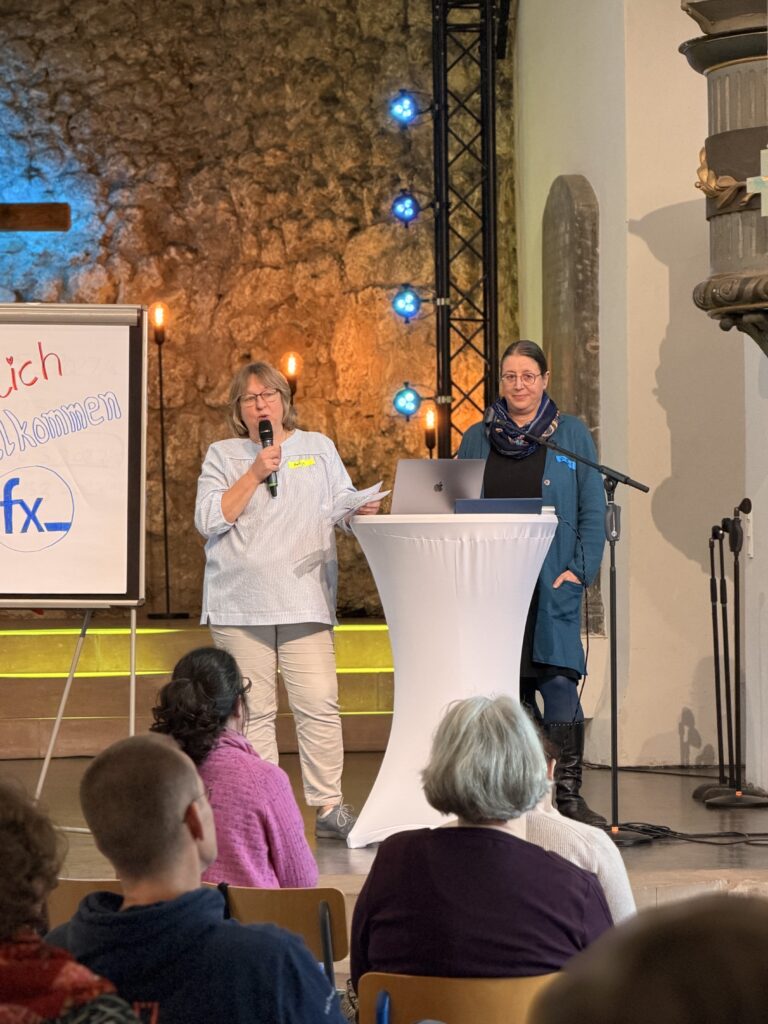 Zwei Frauen stehen an einem Podium und sprechen zu einem Publikum in einem Raum mit Steinwänden; eine hält ein Mikrofon, und auf einer Tafel in der Nähe ist ein Willkommensschild zu sehen.