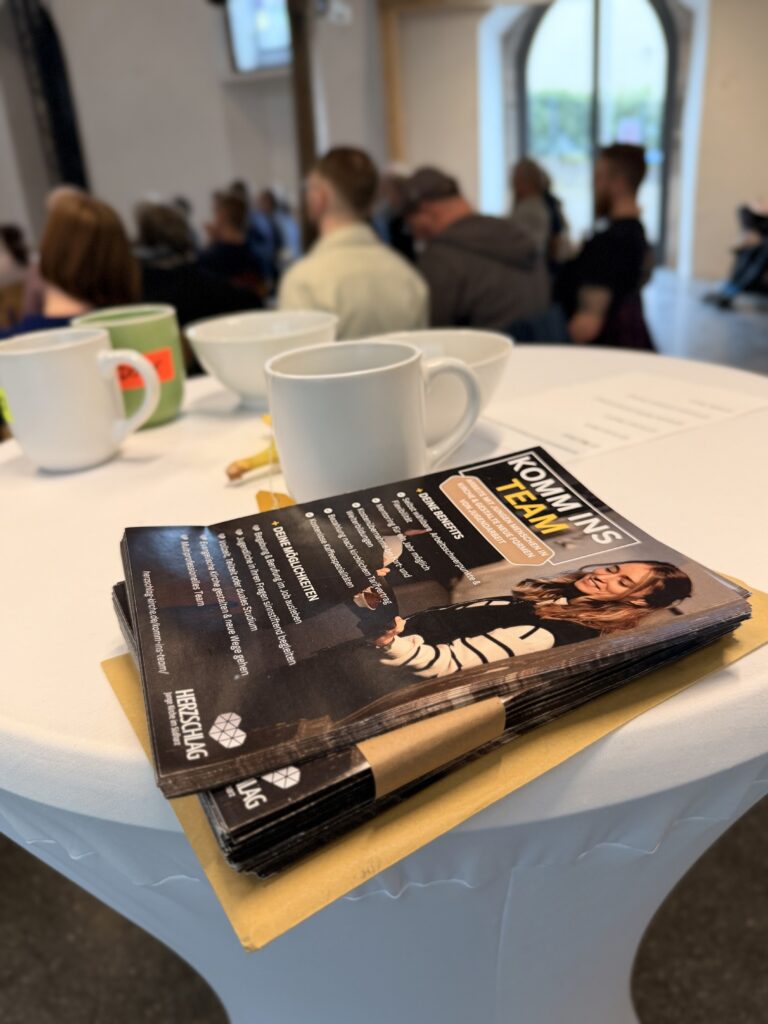 Ein Stapel Zeitschriften und Papiere liegt auf einem runden Tisch mit Kaffeetassen; im unscharfen Hintergrund sind Menschen versammelt und sitzen bei einer Veranstaltung in einem Gebäude.