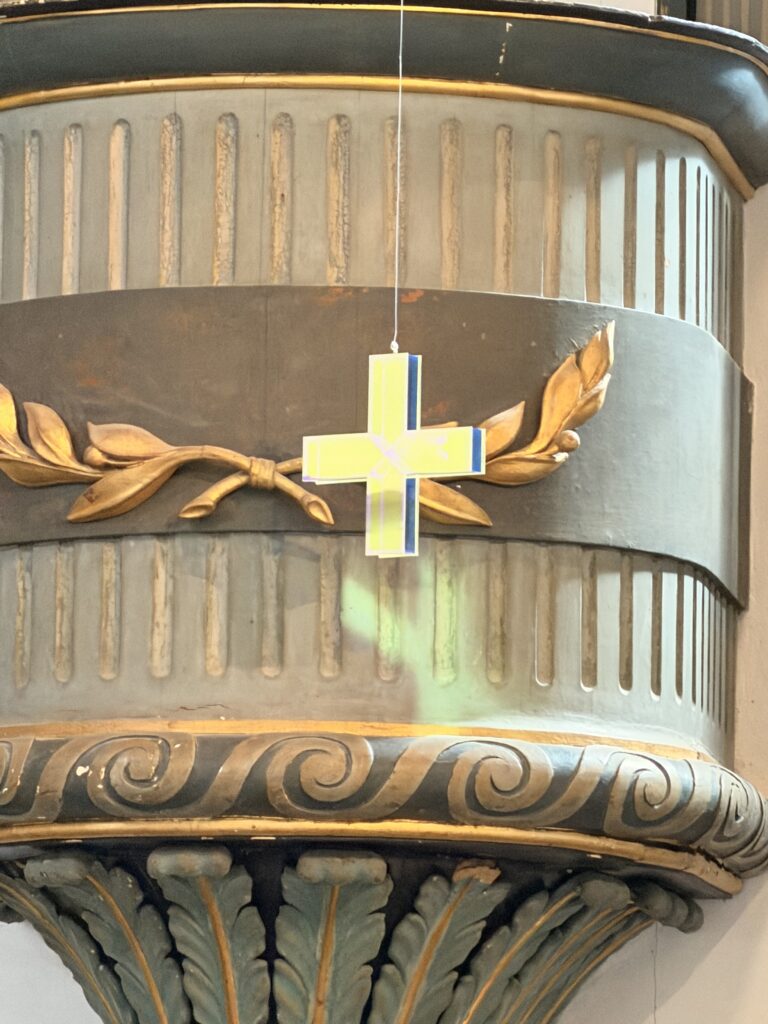 Ein gelbes und blaues Kreuz hängt an einer Schnur vor einer verschnörkelten Säule mit goldenen Lorbeerblättern und vertikalen Rillen.