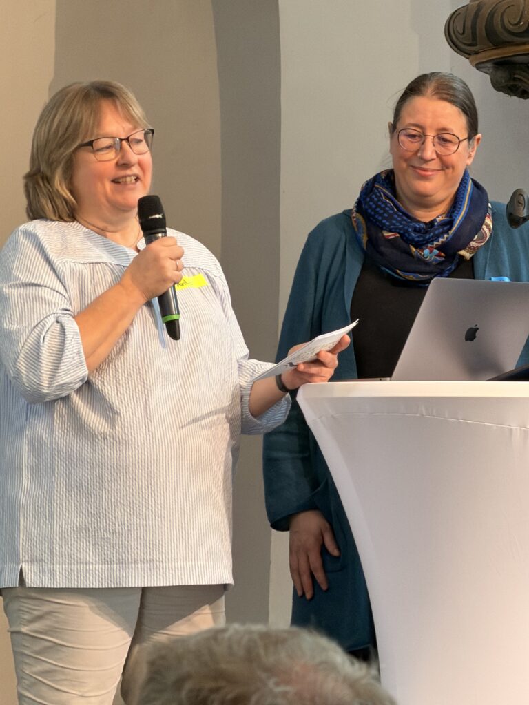Zwei Frauen stehen an einem runden Tisch. Die eine spricht in ein Mikrofon und hält Notizen in der Hand, die andere lächelt und schaut zu, während sie neben einem Laptop steht.