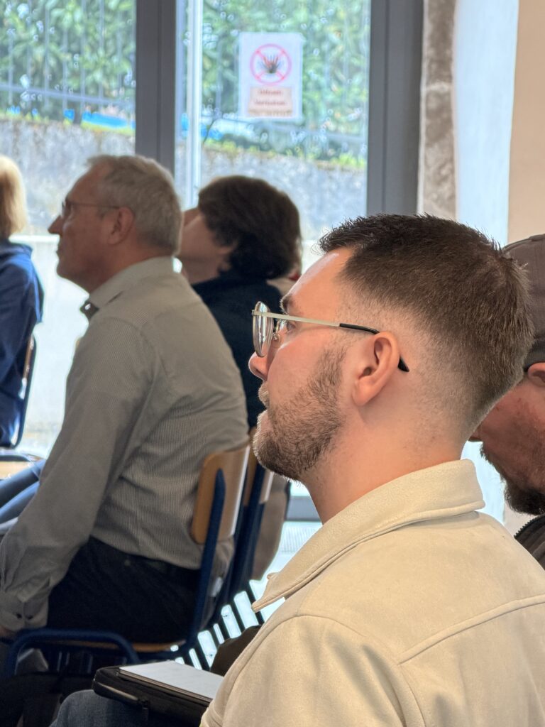 Eine Gruppe von Menschen, die in einem Raum sitzen und aufmerksam zuhören; ein Mann mit Brille und gestutztem Bart steht im Vordergrund.