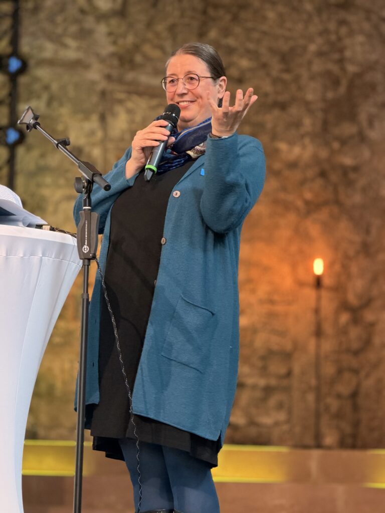 Eine Frau in einem blauen Mantel spricht in ein Mikrofon, während sie mit einer Hand gestikuliert. Sie steht auf einem Podium vor einem verschwommenen, warm beleuchteten Hintergrund.