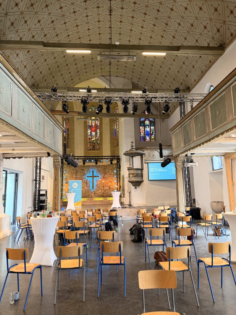 In einem Kirchensaal stehen Stuhlreihen vor einer Bühne mit einem blau beleuchteten Kreuz und Buntglasfenstern, auf der Beleuchtung und Veranstaltungstechnik aufgebaut sind.