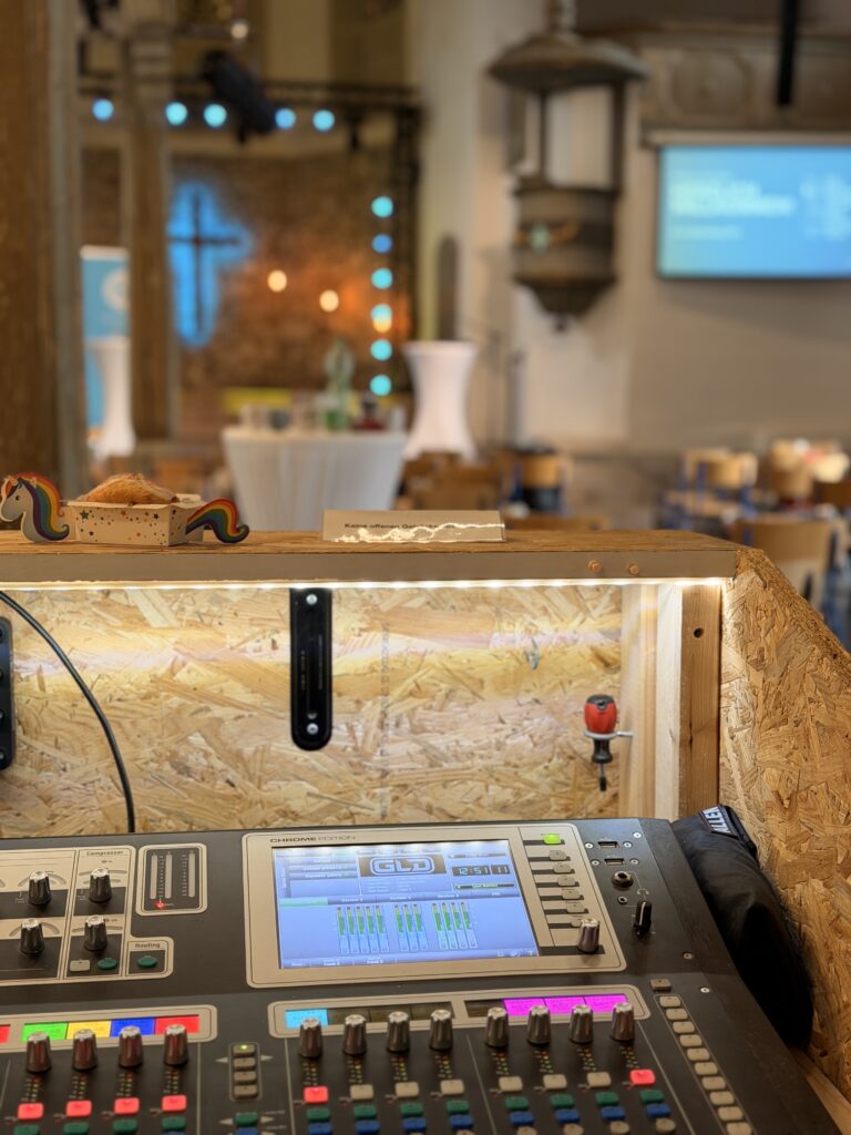 Ein Tonmischpult im Fokus, mit einem unscharfen Hintergrund, der einen Kirchenraum, ein Kreuz, Tische und eine Projektionsfläche mit blauem Text zeigt.