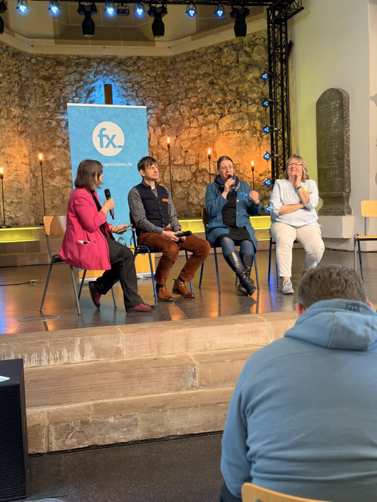 Vier Personen sitzen auf der Bühne vor einer Steinmauer und sprechen bei einer Veranstaltung in Mikrofone, ein Zuschauer steht im Vordergrund.