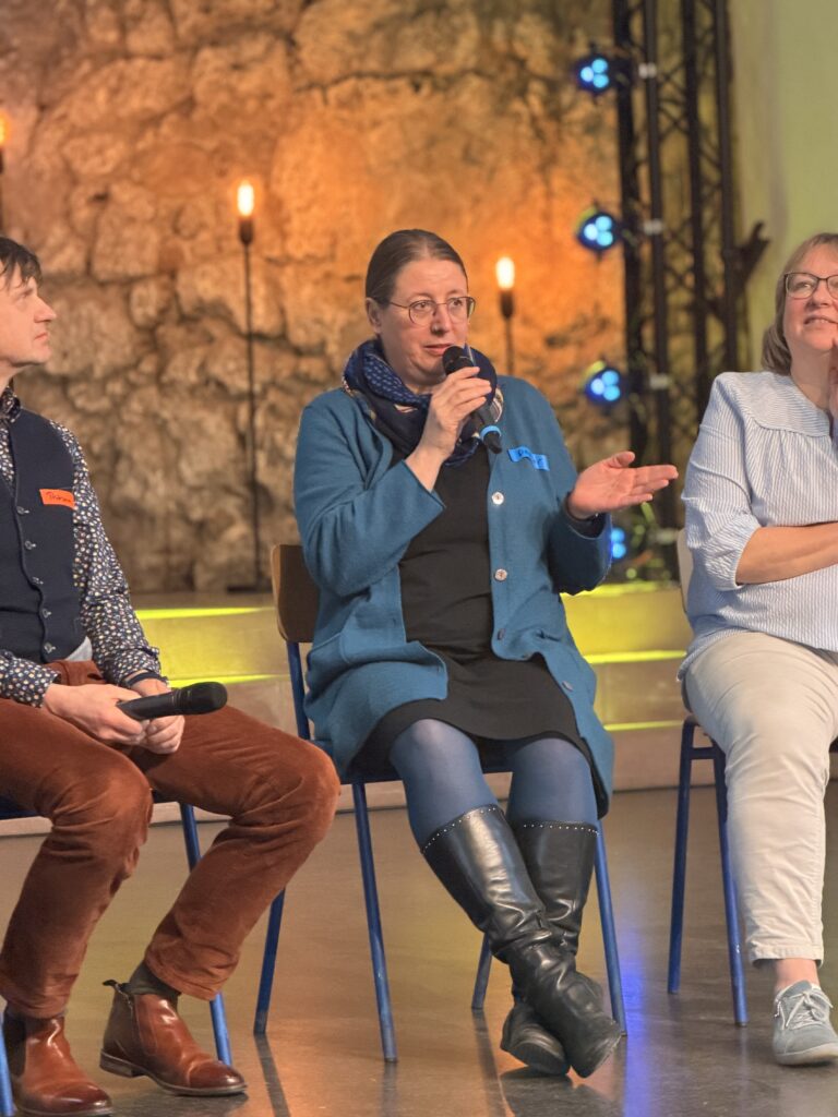 Drei Personen sitzen auf Stühlen; die Frau in der Mitte spricht in ein Mikrofon. Die warme Beleuchtung und die Steinmauer im Hintergrund schaffen eine gemütliche Atmosphäre.