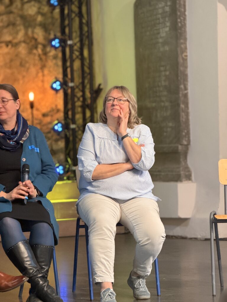 Eine Frau mit Brille sitzt auf einem Stuhl und legt lächelnd die Hand auf ihr Kinn. Sie trägt ein gestreiftes Hemd und eine helle Hose. Andere Menschen sitzen in der Nähe auf einer Bühne.