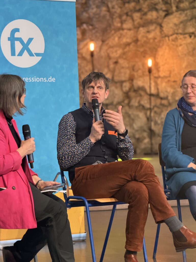 Drei Personen sitzen bei einer Podiumsdiskussion auf der Bühne. Der Mann in der Mitte gestikuliert, während er spricht, während im Hintergrund ein blaues Banner und brennende Kerzen zu sehen sind.