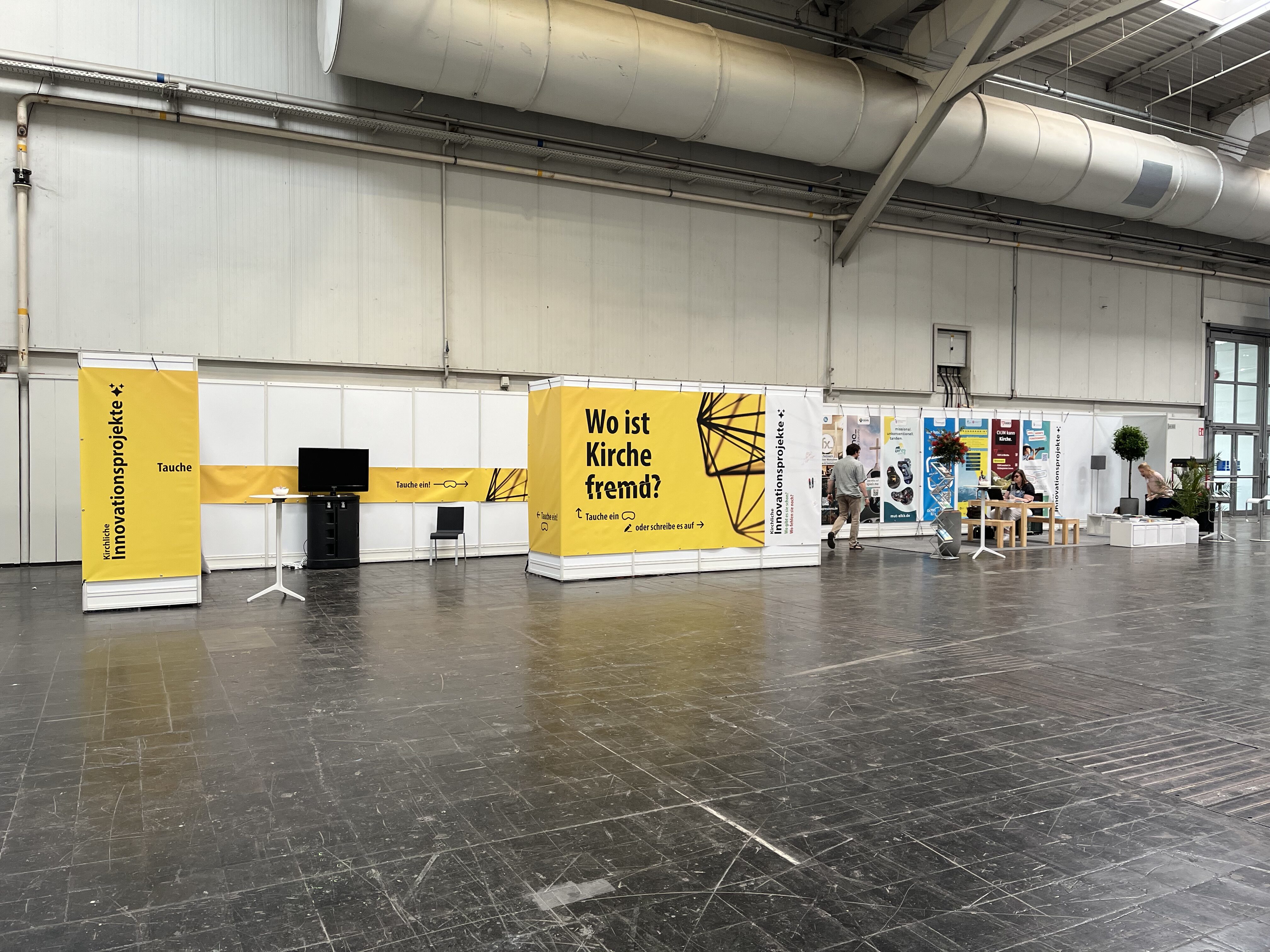 Ein großer überdachter Ausstellungsraum mit gelben Bannern, darunter eines mit der Aufschrift Wo ist Kirche fremd? und Ständen mit Displays, einem Fernseher, Tischen und Menschen, die herumlaufen.