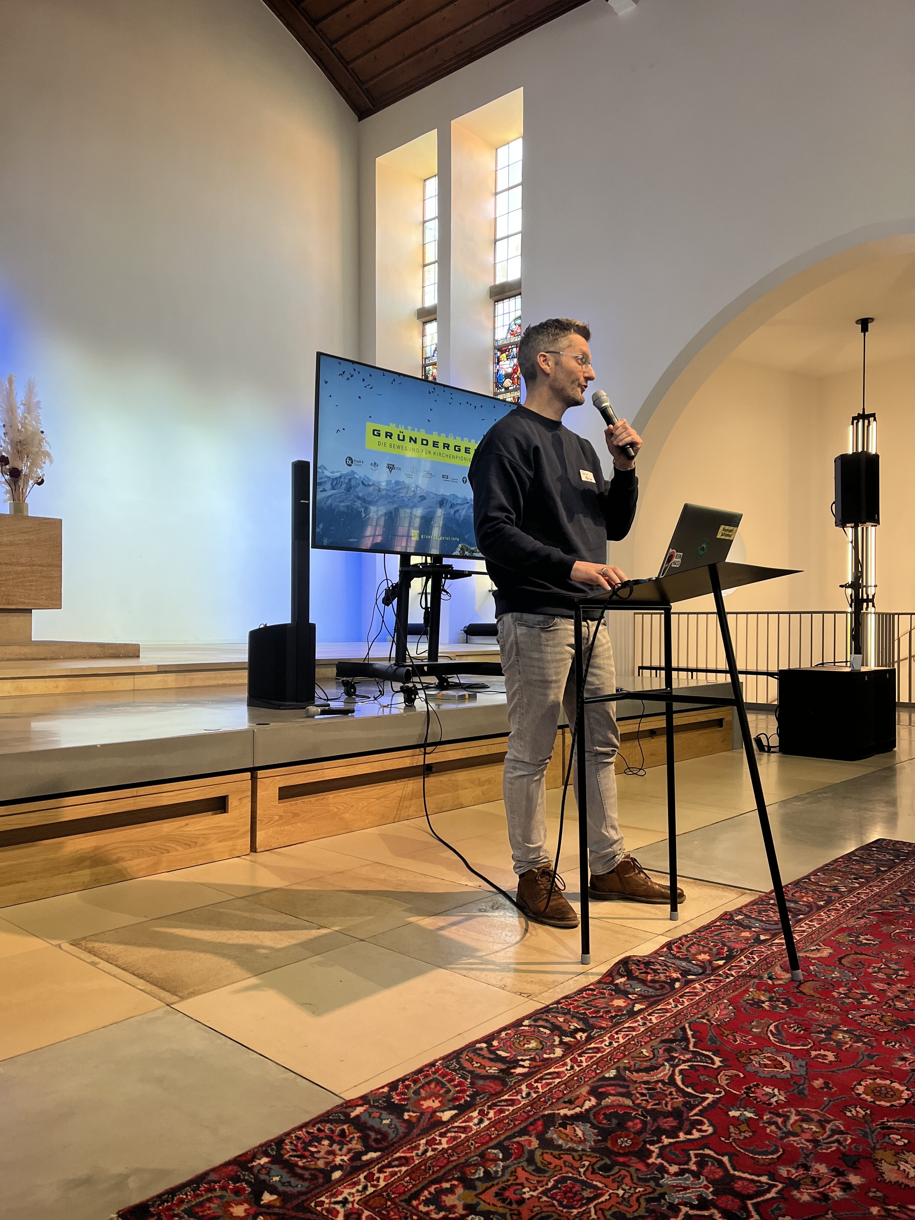 Ein Mann steht an einem Podium mit einem Mikrofon und präsentiert in einer modernen Kirche mit Glasfenstern und einem Fernsehbildschirm mit der Aufschrift "GRONDBOOR".