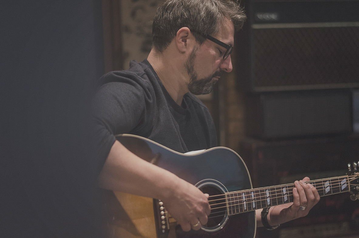 Ein Mann mit Brille und Bart spielt drinnen auf einer akustischen Gitarre und konzentriert sich auf seine Musik.