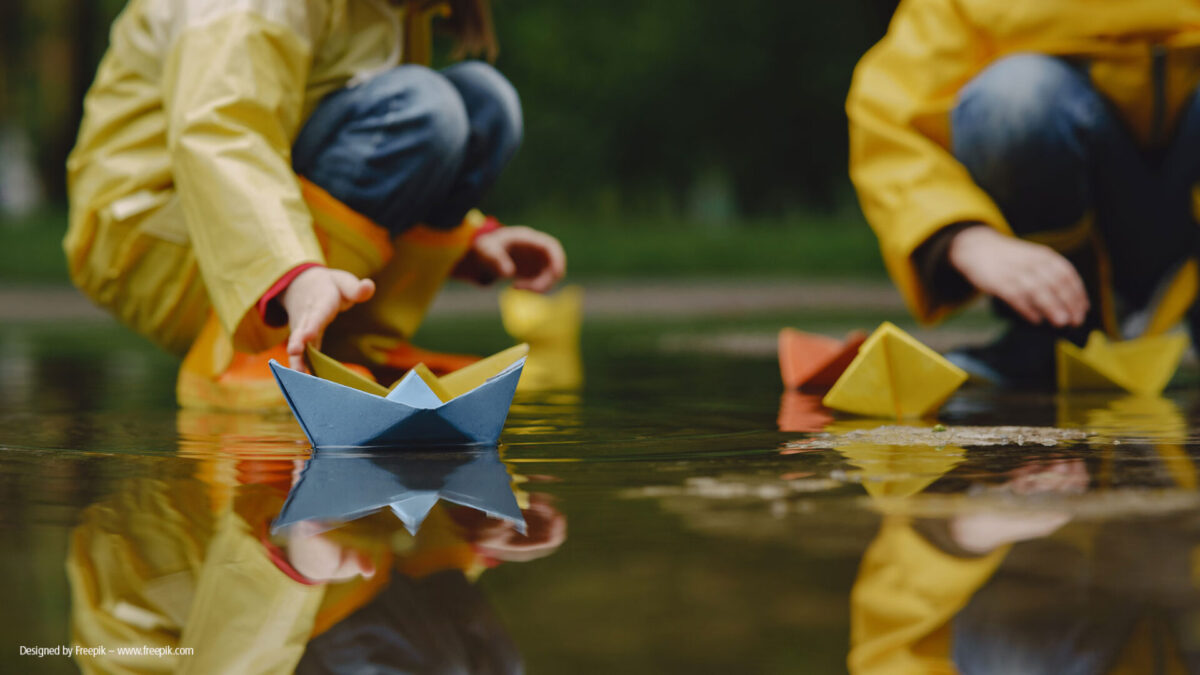 Zwei Kinder in gelben Regenmänteln spielen an einer Pfütze und lassen bunte Papierboote auf das Wasser gleiten, wobei die Bäume im Hintergrund verschwimmen.