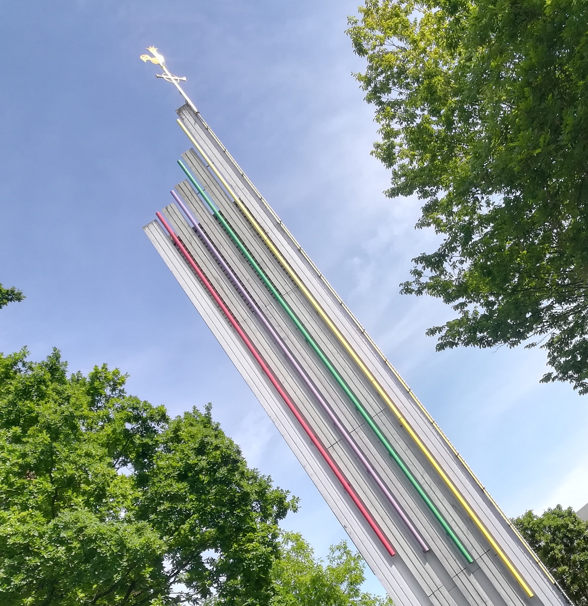 Hohe Metallskulptur mit bunten vertikalen Linien und einer goldenen Figur an der Spitze, umgeben von grünen Bäumen unter einem blauen Himmel.